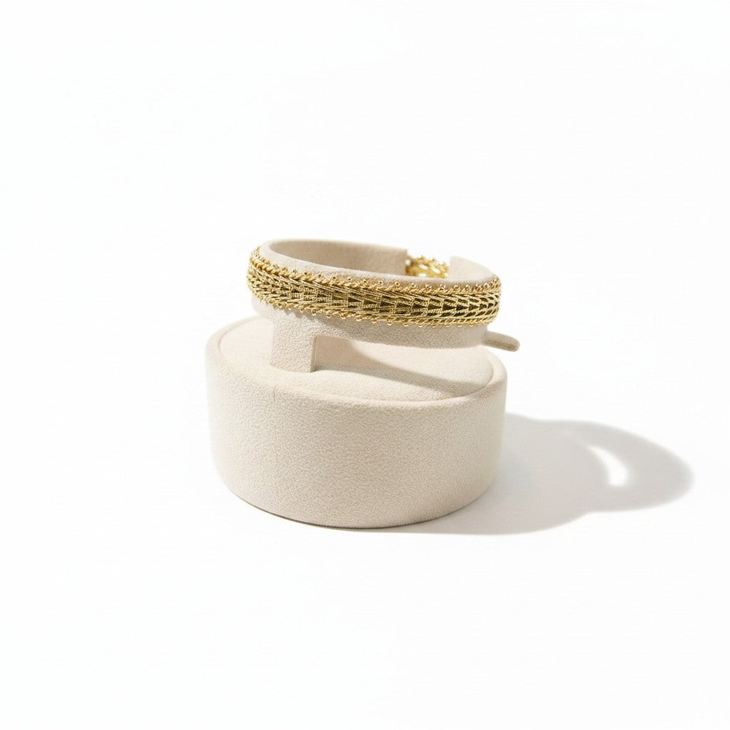 ANIBA JEWELS gold gourmette bracelet with intricate woven pattern and beaded edges, displayed on a beige stand.
